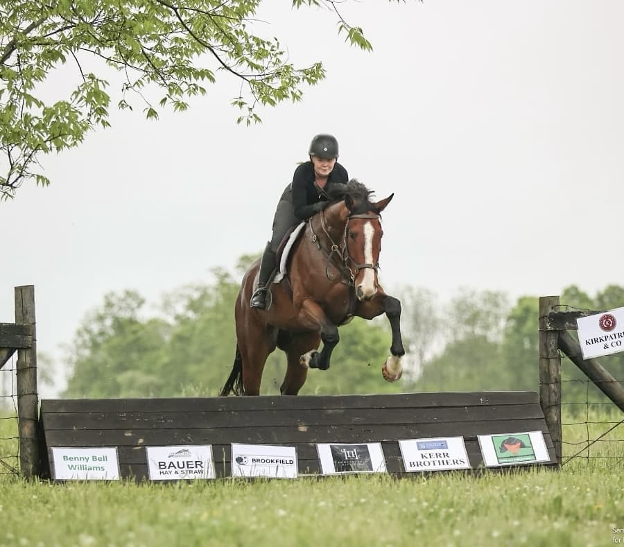 Mackenzie riding Hawksbridge, a bay sport horse, clearing a sponsored cross-country oxer